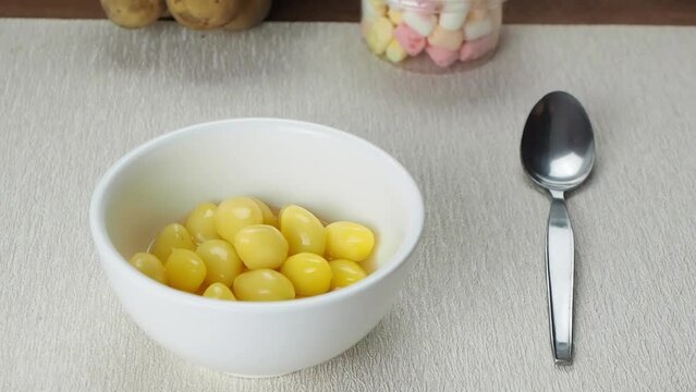 Pouring gingko seeds with syrup and ice cubes into white ceramic bowl with metal spoon on table mat on wooden table.