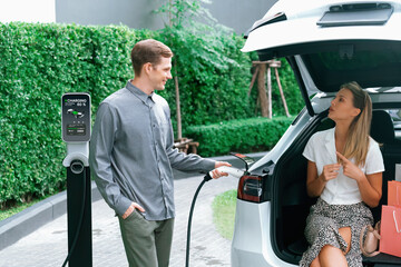 Young couple travel with EV electric car charging in green sustainable city outdoor garden in...
