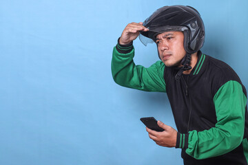 Asian man with helmet wear green jacket, holding smartphone and looking far away isolated on blue...