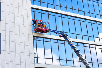 External glass facade window washers, while in a construction lifting cradle, clean the glass facade of a building on a clear day. - 668008929