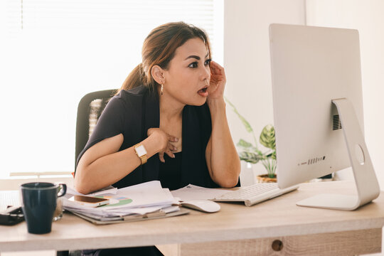 Adult Asian business woman sitting on desk and looking at computer screen with shocked surprise expression