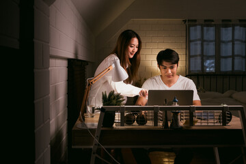 Asian couple working late from home with a laptop on the table in the dark bedroom. Husband and wife discuss and brainstorm together at night before the business work deadline. Night working concept.