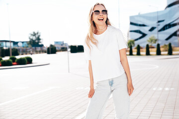 Portrait of beautiful smiling model. Female dressed in summer hipster white T-shirt and jeans. Posing  in the street at sunny day. Funny and positive woman having fun outdoors. In sunglasses