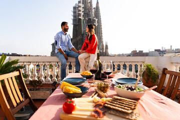 Multiracial beautiful happy couple of lovers dating on rooftop balcony at Sagrada Familia, Barcelona - Multiethnic people having romantic aperitif dinner on a terrace with city view  - obrazy, fototapety, plakaty