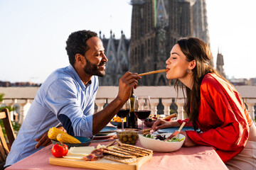Multiracial beautiful happy couple of lovers dating on rooftop balcony at Sagrada Familia, Barcelona - Multiethnic people having romantic aperitif dinner on a terrace with city view 