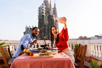 Schilderijen op glas Multiracial beautiful happy couple of lovers dating on rooftop balcony at Sagrada Familia, Barcelona - Multiethnic people having romantic aperitif dinner on a terrace with city view  © oneinchpunch