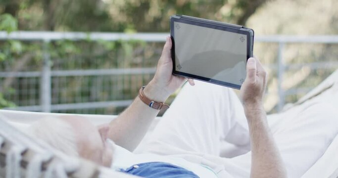 Caucasian middle aged man using tablet relaxing in hammock on terrace in nature, slow motion