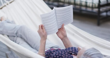 Caucasian middle aged woman reading book relaxing in hammock on terrace in nature, slow motion