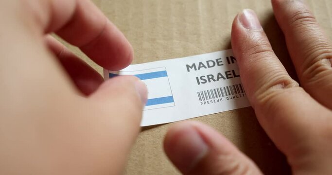 Hands applying MADE IN ISRAEL flag label on a shipping box with product premium quality barcode. Manufacturing and delivery. Product factory import and export.
