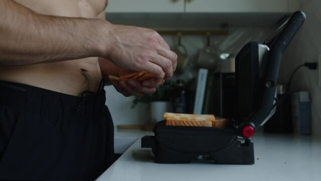 Man walks up to a panini grill press and places marble cheese slices onto two pieces of white bread, preparing to make grilled cheese on his white countertop.