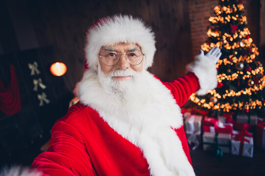 Photo of funky good mood santa claus dressed red eyewear recording video vlogging showing arm tree indoors christmas north pole home