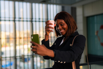 Fototapeta premium In the mall's hallway, the formally dressed black business woman engages in a video call, raising her coffee in a toast to the person she's speaking with.
