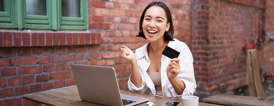 Enthusiastic asian girl sitting with credit card and laptop, paying contactless, shopping online via computer