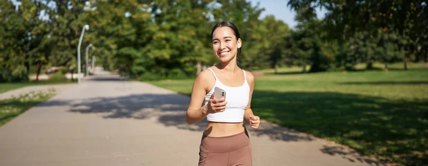 Poster Happy smiling asian woman jogging in park. Healthy young female runner doing workout outdoors, running on streets © Mix and Match Studio