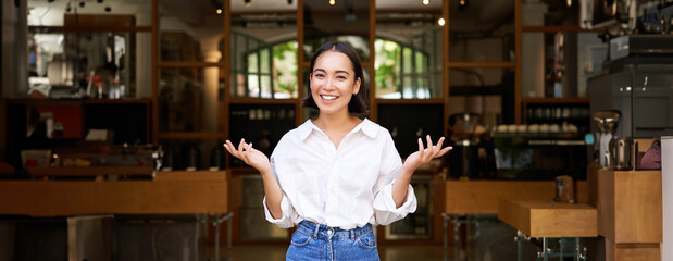 Enthusiastic young businesswoman, asian girl showing her business, raising hands up and smiling, standing in front of restaurant - obrazy, fototapety, plakaty