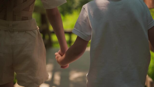 Little girl holds toddler boy hand walking together along overpass in forest closeup. Children enjoy summer and friendship at countryside nature