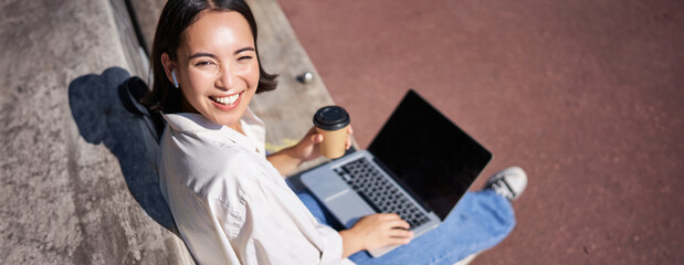 Beautiful young asian female student, sitting with laptop on bench in park, drinking takeaway coffee and listening music, working remotely, studying outdoors