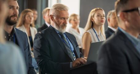 Senior Male Top Manager Sitting In Crowd And Using Laptop Computer At International Technology Conference. Caucasian Man Listening To Keynote Presentation About Innovative Software Solution.