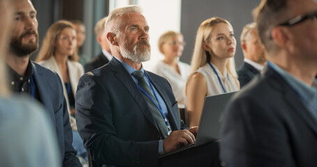 Senior Male Investor Sitting In Crowd And Using Laptop Computer At International Technology...