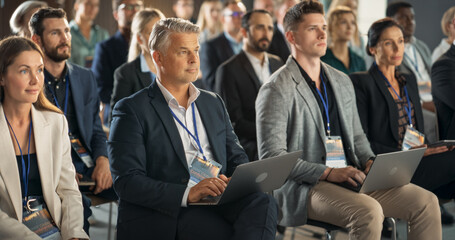 Caucasian Male Pharmaceutical CEO Sitting In Diverse Crowd At Medical Summit And Using Laptop Computer. Successful Man Listening To Presentations About New Developments In Biopharma and Medicine.