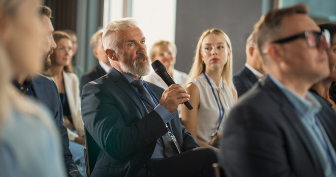 Senior Male Investor Sitting In Crowd And Using Microphone To Ask A Question At International Technology Conference. Caucasian Man Listening To Keynote Presentation About New Startup Company Service.