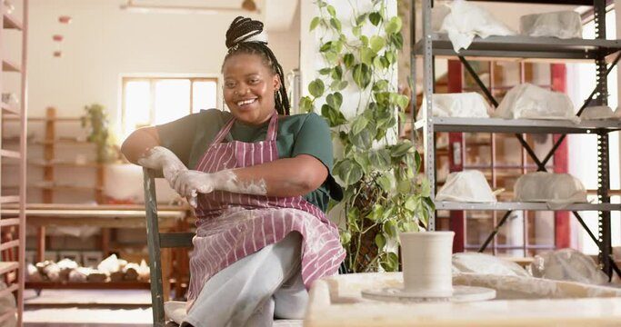 Happy african american female potter sitting and smiling in pottery studio, slow motion