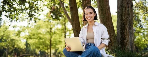 Portrait of asian girl with laptop, sitting near tree in park on sunny summer day, working on remote, e-learning with computer