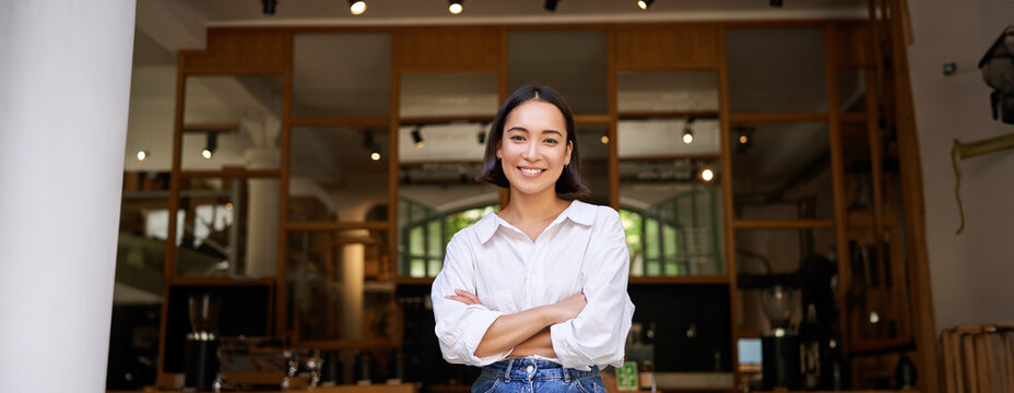 Portrait of confident young asian woman, business owner, cross arms on chest, looking pleased, standing in front of cafe