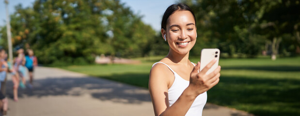 Happy asian fitness girl running and checking her stats, daily goals on smartphone app. Young woman...