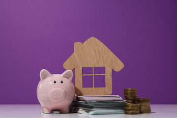 Wooden house, money and piggy bank on purple background