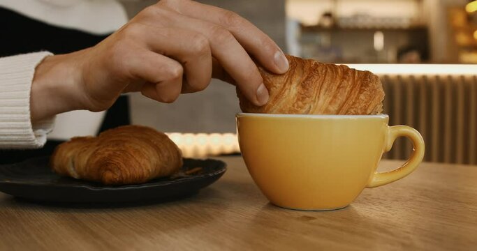 Close-up of female hand dunking half of freshly baked croissant into a cup of black coffee, drops falling. Eating in a bakery. Lifestyle horizontal video.