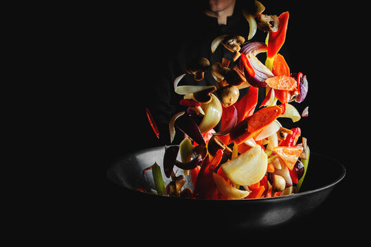 Closeup of chef throwing vegetable mix from wok pan on black background.