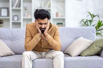 Sad depressed man sitting at home on sofa in living room, hopelessly lonely.