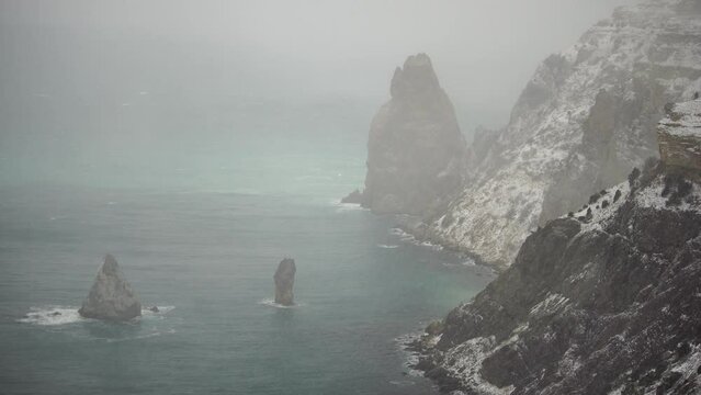 Snow covered rocky cliffs over sea. Winter landscape over the sea bay. Juniper tree under snow. Concept of winter holidays, travel and recreation. Weather and Climate Change.