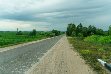 Fototapeta na wymiar An empty roadway outside the city passes by agricultural fields. A highway with a new asphalt surface on the background of fluffy clouds. Paved road on a sunny day without cars.