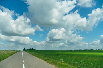 An empty roadway outside the city passes by agricultural fields. A highway with a new asphalt surface on the background of fluffy clouds. Paved road on a sunny day without cars.