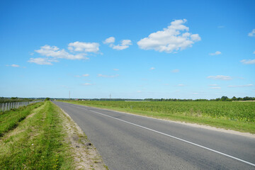 An empty roadway outside the city passes by agricultural fields. A highway with a new asphalt surface on the background of fluffy clouds. Paved road on a sunny day without cars.
