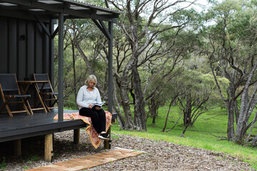 Woman reading a book on the deck of a tiny house and relaxing in the Australian bush - obrazy, fototapety, plakaty
