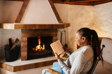 Senior African woman reading book while warming in front of fireplace in her house