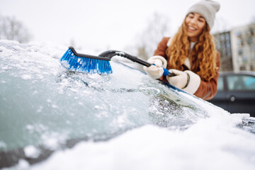 Time to clear the snow! A young woman cleans snow from a car with a brush. Winter car concept.