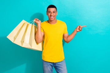 Portrait of cheerful funny man hand hold boutique bags indicate finger empty space isolated on...