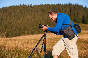 Professional photographer taking pictures in mountains. Man focusing his camera to take nice photo. Side view of young male adjusting camera on background of hills.