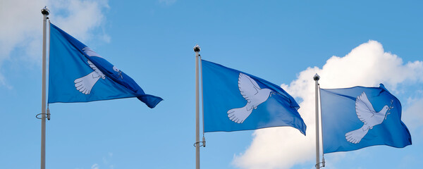 Three blue peace flags in a row printed with a dove with an olive branch in its beak. The dove with...