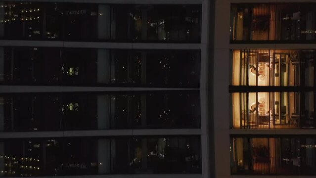 Ascending fly along high rise apartment building at night. People in flats with lighted windows. Abstract computer effect digital composed footage