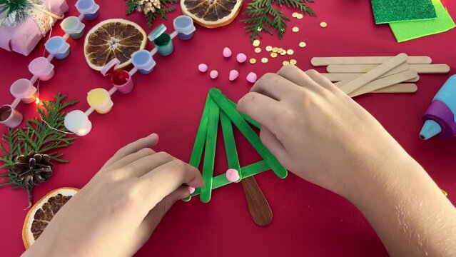 how to make christmas tree from ice cream sticks. Child decorates with pink balls New year handmade craft tree. Concept of DIY and kid's creativity.