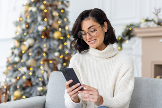 A young female student is texting on the phone, reading the news. He sits on the sofa near the Christmas tree and looks at the screen with a smile