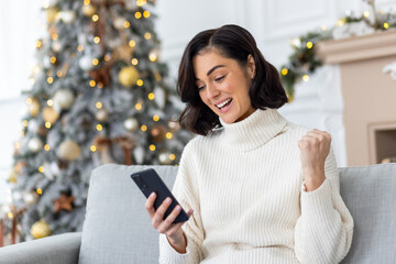 Happy young woman sitting at home on sofa near Christmas tree and using mobile phone, happy with success and good news for New Year holidays, showing victory gesture with hand - obrazy, fototapety, plakaty