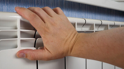 Unrecognizable man's hand check for heat and touch central heating system. White metal heating iron household radiator inside room. Budget in crisis concept. House in winter
