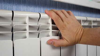 Unrecognizable man's hand check for heat and touch central heating system. White metal heating iron household radiator inside room. Budget in crisis concept. House in winter