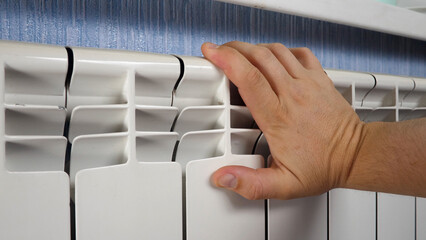 Unrecognizable man's hand check for heat and touch central heating system. White metal heating iron household radiator inside room. Budget in crisis concept. House in winter